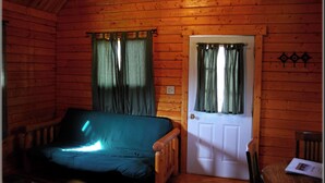 A wooden cabin with a single bed, a door with curtains, and a small table with a book. - Beautiful Romantic River front cabin with mountain views,fantastic location! (Luray)