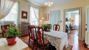 A dining room with a lace tablecloth, wooden chairs, potted plants, and a radiator.
