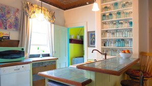 A kitchen with a built-in island, a window with curtains, a shelf with jars, and a chair.