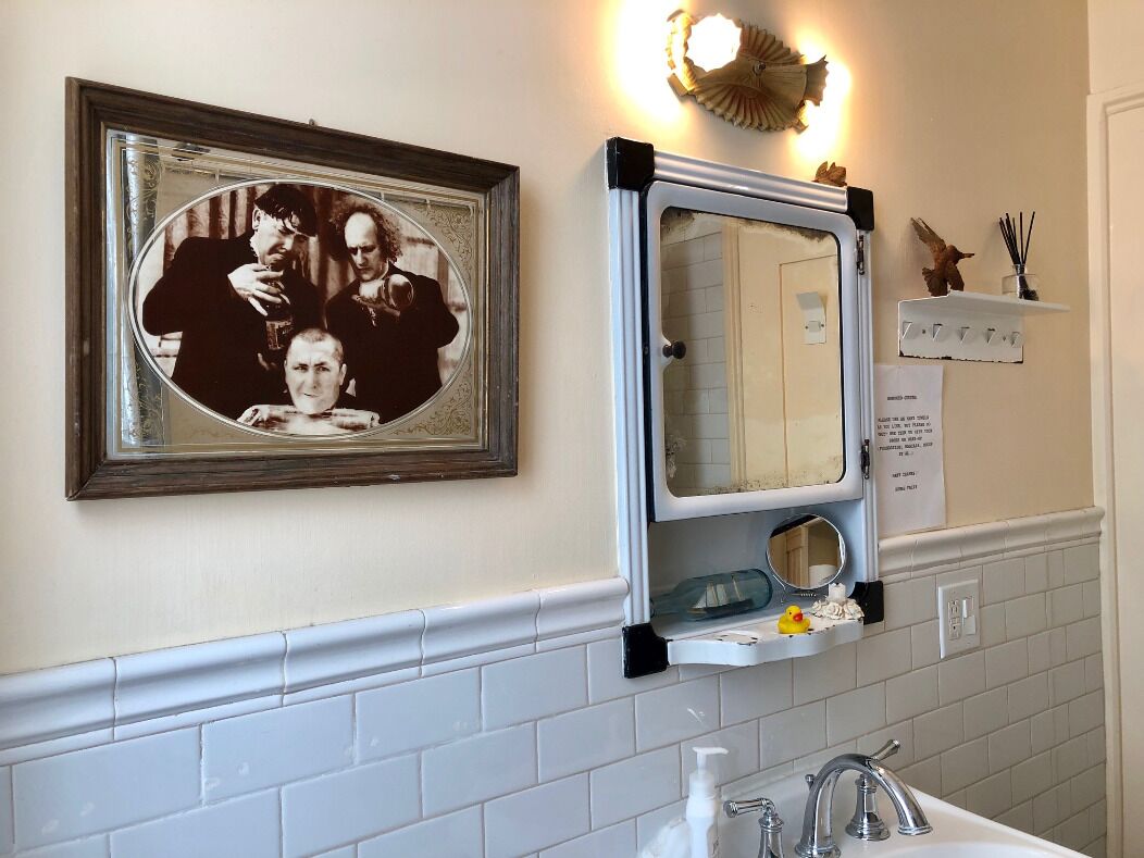 Un baño con pared de azulejos blancos, una fotografía enmarcada, un espejo y un estante pequeño con artículos de aseo.