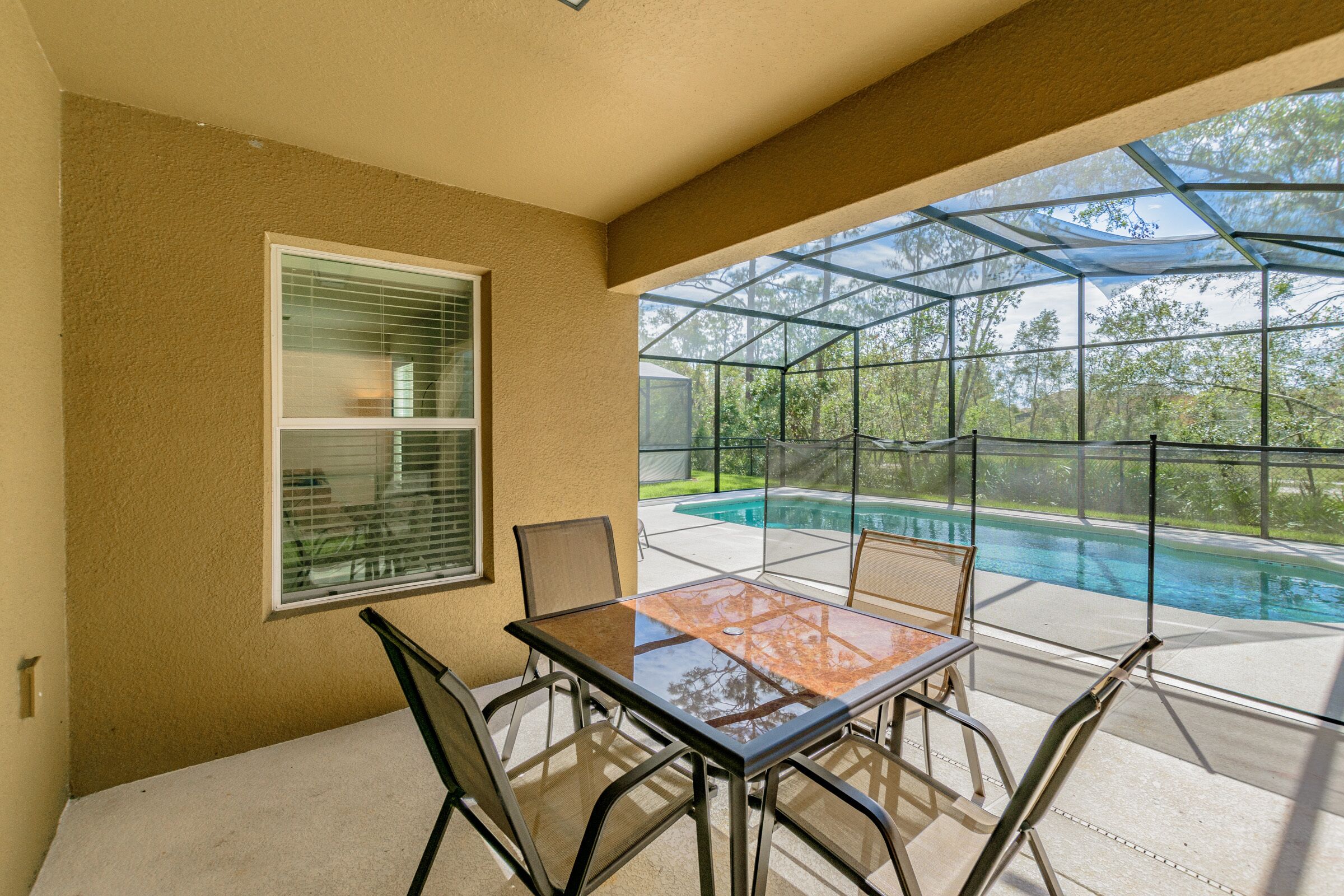 A covered patio with a glass table and chairs, overlooking a swimming pool and trees.