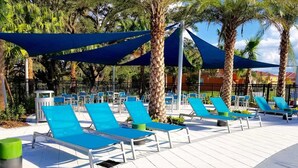A poolside area with blue lounge chairs and umbrellas, surrounded by palm trees and a tiled walkway.