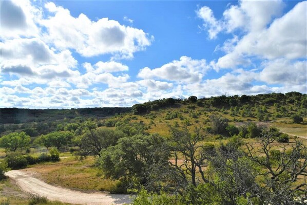 A rural landscape with a dirt road, scattered trees, and a hilly terrain under a partly cloudy sky.