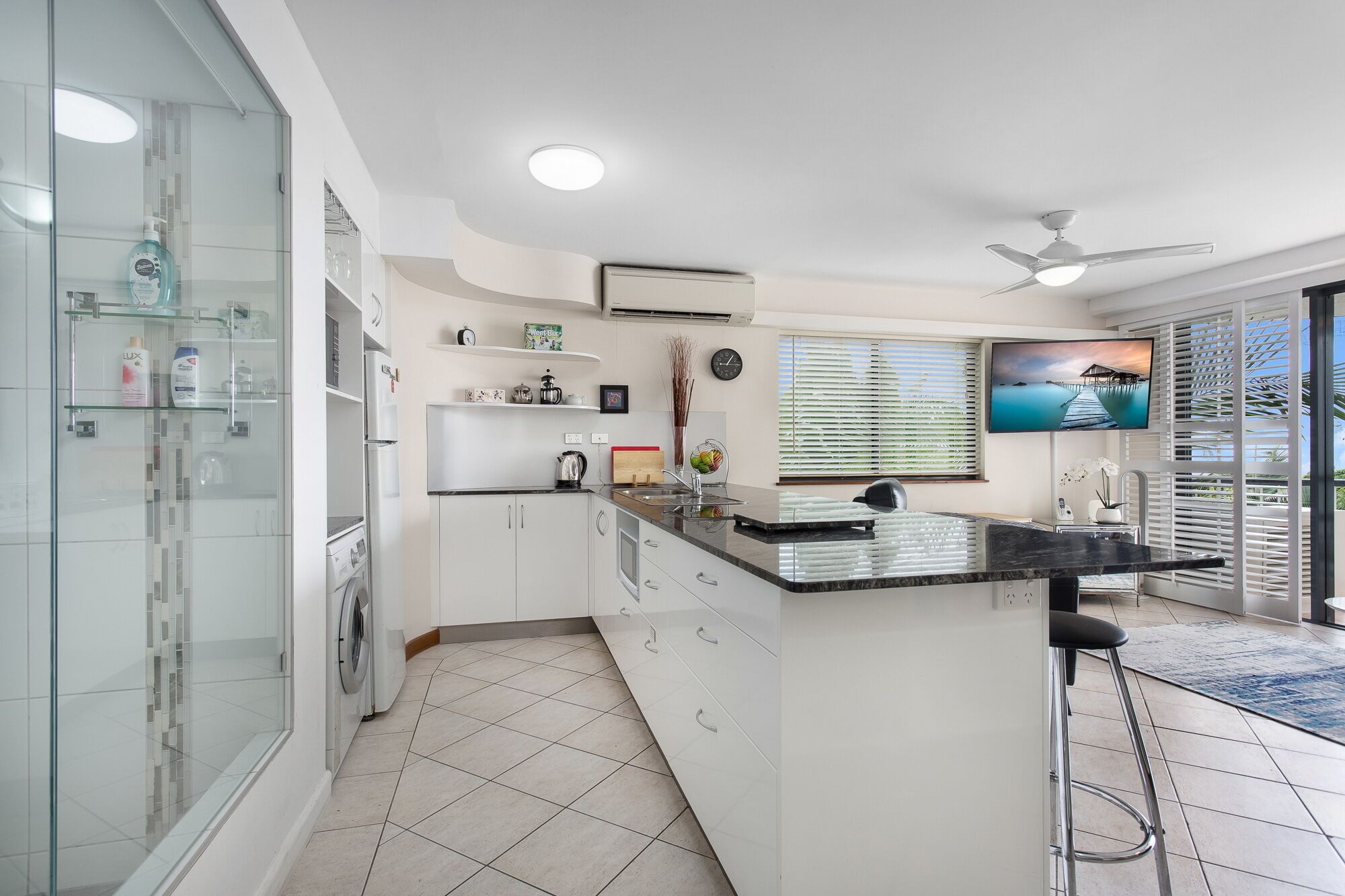 A modern kitchen with a central island, a built-in washing machine, and a large window with blinds.