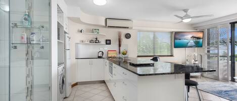 A modern kitchen with a central island, a built-in washing machine, and a large window with blinds.