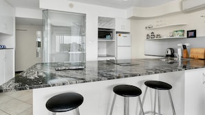 A modern kitchen with a marble countertop, three black bar stools, white cabinets, and a glass shower enclosure. - Best Location in Cairns - Ocean Views (Cairns)