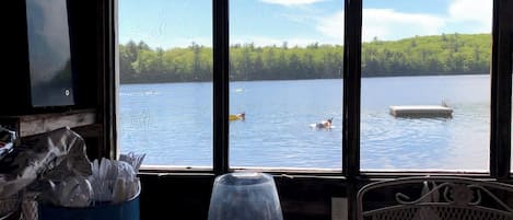 A room with a large window overlooking a lake, a table with a glass, and a chair.