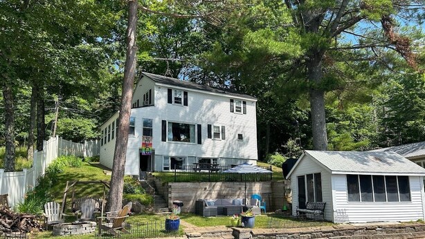 A two-story house with a dock extending into a body of water, surrounded by trees and a fenced yard.