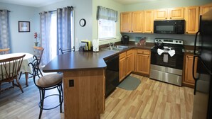 A kitchen with wooden cabinets, a stainless steel refrigerator, a microwave, and a stove. There is a dining table with chairs and a window with curtains.