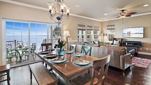 A spacious living area with a dining table set for four, a large window overlooking a body of water, a ceiling fan, and a television mounted on the wall. - Stunning Waterfront Villa w Incredible Views - Htd Pool, Spa, Dock, Beach, Gm Rm (Mary Esther)