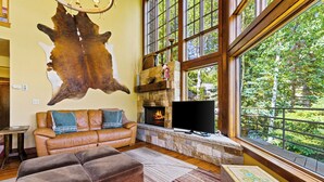 Ein gemütliches Wohnzimmer mit Ledersofa, Hocker, Kamin aus Stein und großen Fenstern mit Blick auf einen Wald.