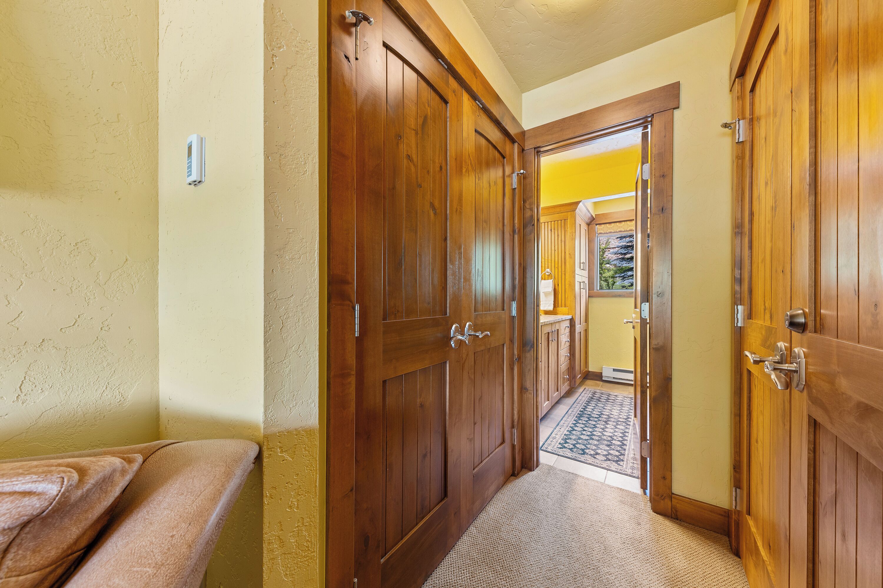 Un couloir avec des portes en bois, un banc et un tapis menant à une pièce dont un mur est jaune et qui possède une fenêtre.