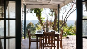 A covered outdoor dining area with a table set for two, surrounded by wooden chairs, overlooking a scenic landscape with trees and mountains. - Gladville- old world charm with stunning views (Daylesford)