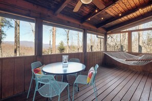 Eine Holzterrasse mit einem runden Tisch, Stühlen, einer Hängematte und großen Fenstern mit Blick auf einen bewaldeten Bereich.