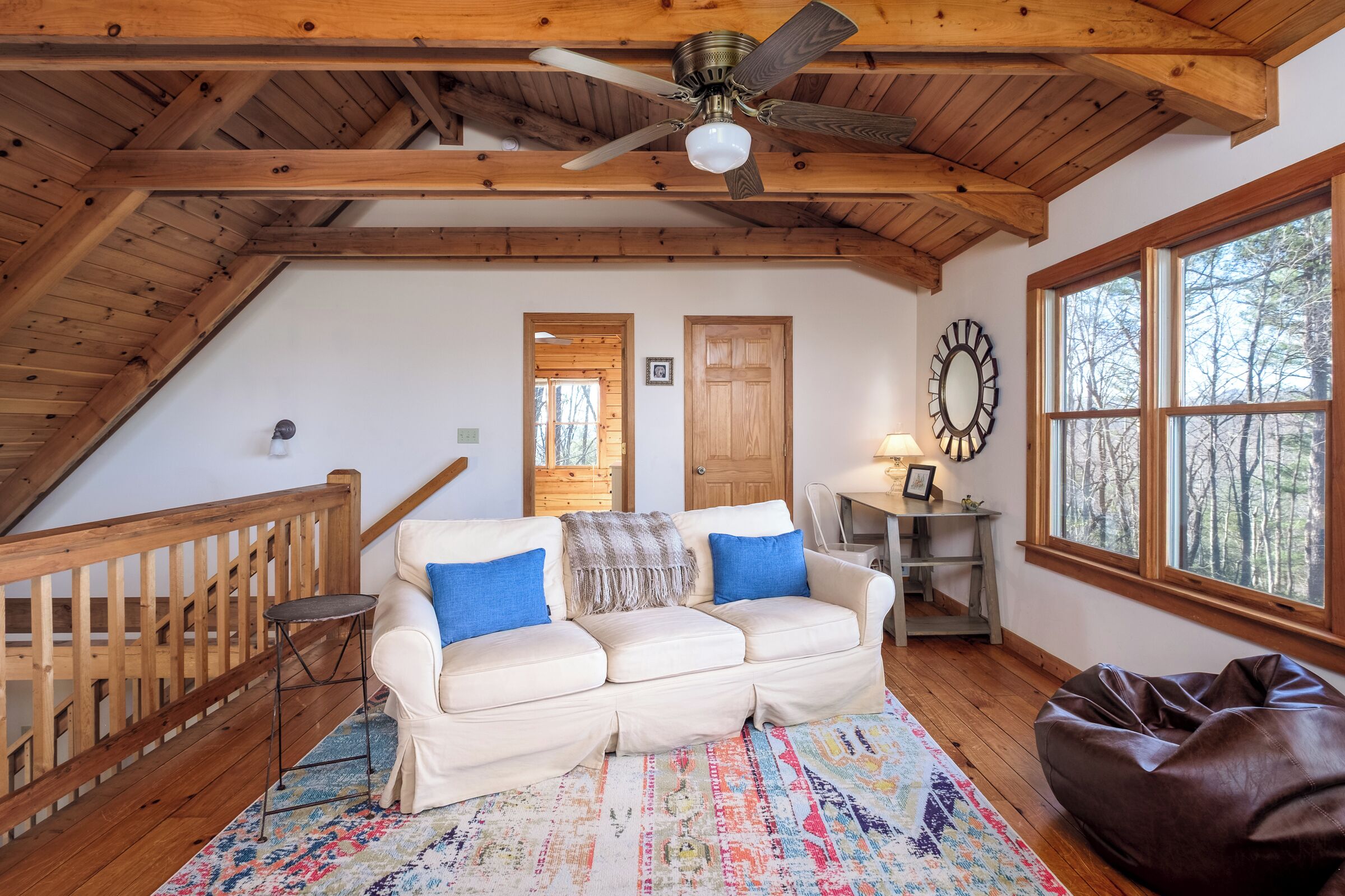 Une chambre à coucher confortable, aménagée en mezzanine, avec un plafond en bois, un canapé blanc agrémenté de coussins bleus, un tapis à motifs et une grande fenêtre donnant sur une forêt.