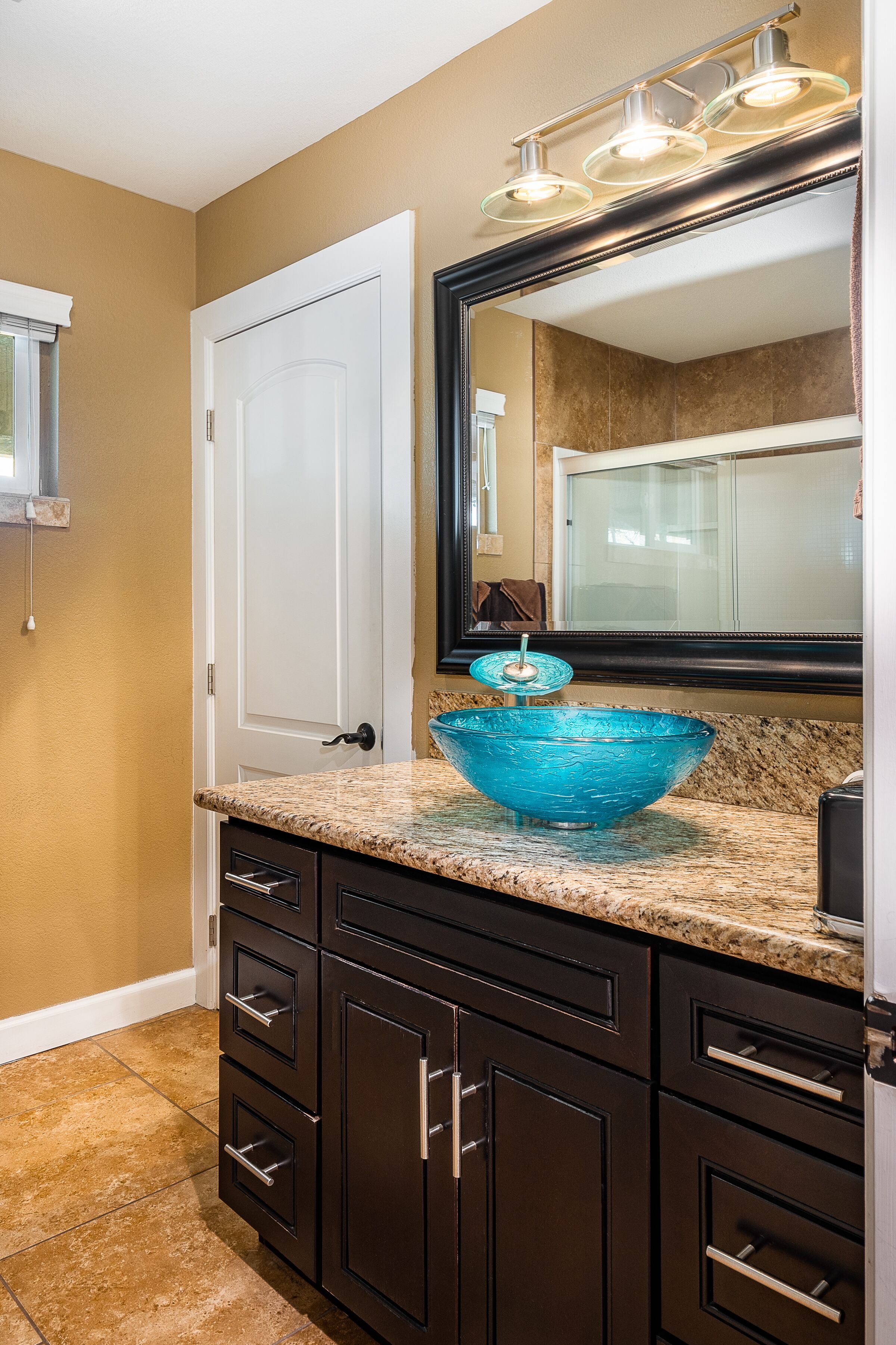 A bathroom with a large mirror, a blue glass bowl sink, dark cabinets, and a beige wall.