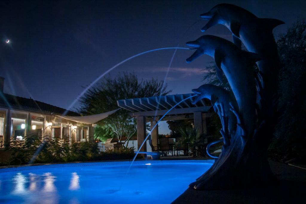 A nighttime scene with a swimming pool, illuminated by artificial lights. There is a sculpture of dolphins and a covered area with lights.