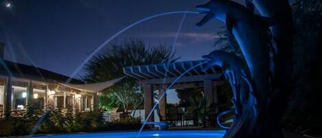 A nighttime scene with a swimming pool, illuminated by artificial lights. There is a sculpture of dolphins and a covered area with lights.