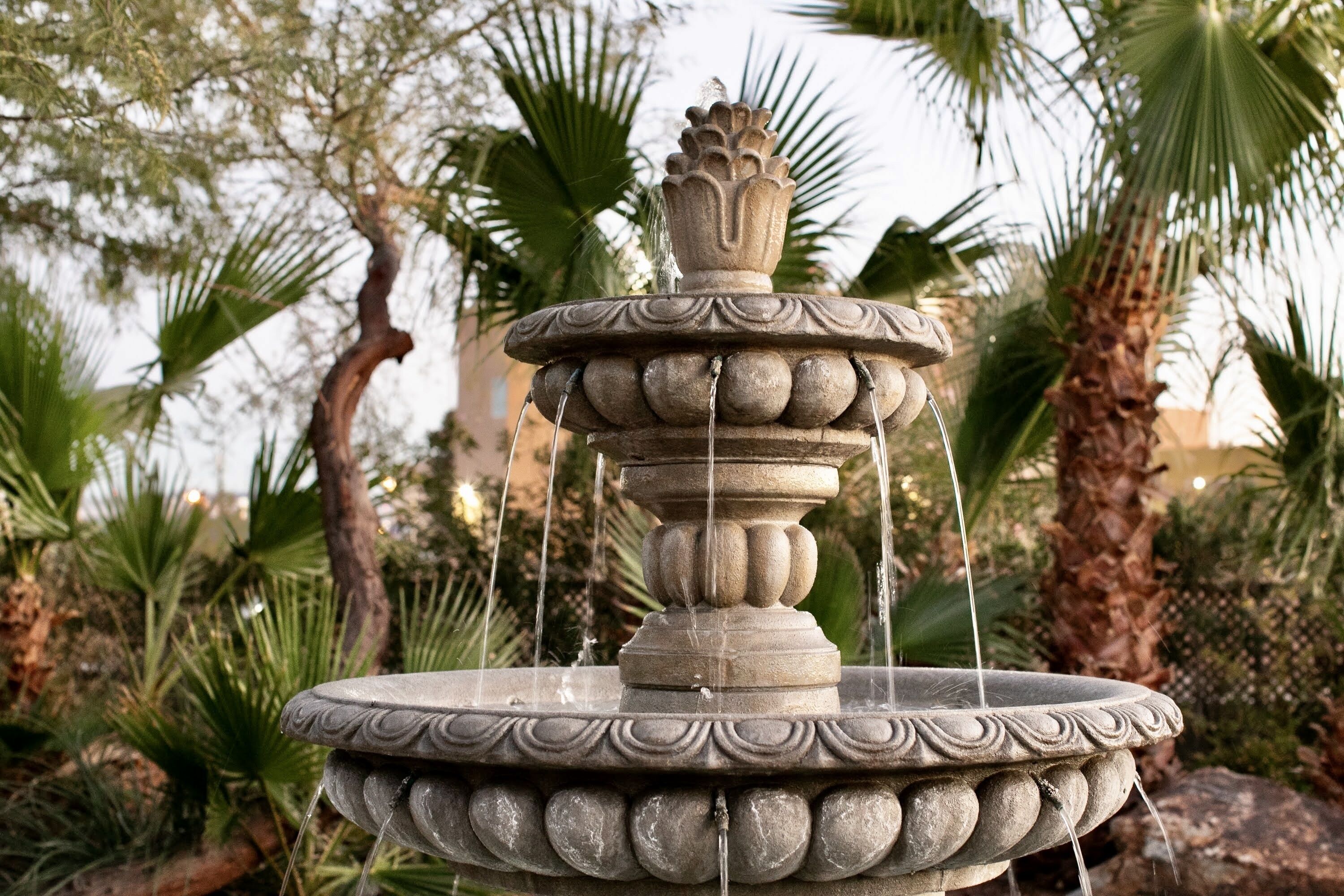 Une fontaine en pierre à plusieurs niveaux, dont l’eau jaillit de l’étage supérieur, le tout entouré de palmiers et d’autres plantes vertes.