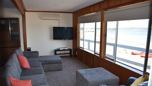 Un salon doté d’un canapé d’angle gris, de poufs, d’un téléviseur à écran plat et d’une grande fenêtre donnant sur la plage.