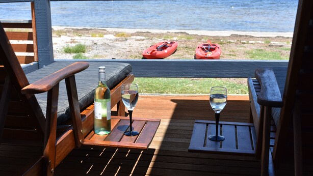 Une terrasse en bois avec vue sur la mer, deux kayaks et une bouteille de vin sur une table.