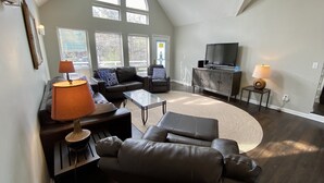 A spacious living room with a high ceiling, large windows, a ceiling fan, a flat-screen TV, and a fireplace. - Big Kitchen, Two Masters, Hot Tub, Short Walk from Croatan Beach (Virginia Beach)