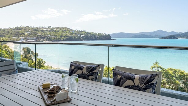 A balcony with a table set for two, overlooking a coastal view with a mountain in the distance. - Frangipani Lodge 207, Beachfront Apartment on Hamilton Island (Hamilton Island)