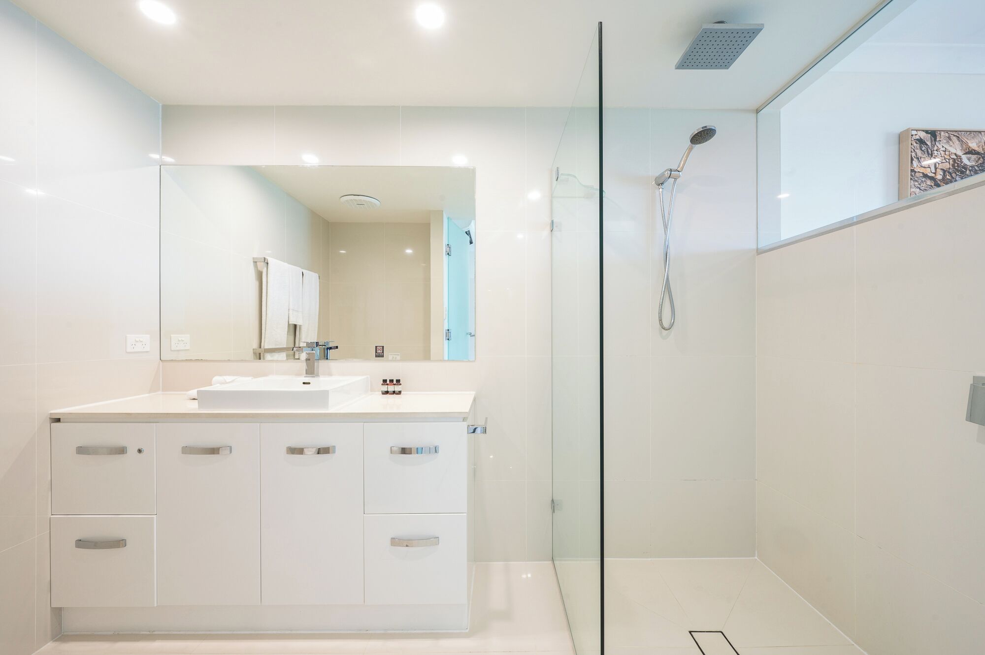 A modern bathroom with a glass-enclosed shower, a large mirror, and a white vanity with multiple drawers.