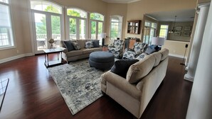A spacious living room with a large sectional sofa, a coffee table, and a rug. There are multiple windows providing natural light and a view of the outdoors. - Grand-Escapes on Norris Lake-Available May July3-10, July 17-24, July 31-Aug 14. (La Follette)