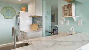 A kitchen with a washing machine, a sink, a marble countertop, and a wall-mounted shelf with decorative items. - Sea Colony Oceanfront 1 Bedroom Condo Beautiful View Sept 21 -25 Super Special  (Bethany Beach)