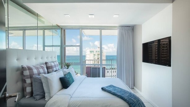 A modern bedroom with a large bed, a view of the sea, and a cityscape through large windows. - Ocean View  19 floor  STUDIO Apt In the Condado Area NO PARKING (San Juan)