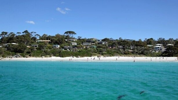 Une zone côtière dotée d’une plage, d’eaux turquoise et limpides, et d’une rangée de maisons adossées à une colline.