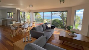 Un vaste espace de vie comprenant un grand canapé gris, une table à manger en bois et des chaises. De grandes fenêtres offrent une vue sur la mer et la verdure.
