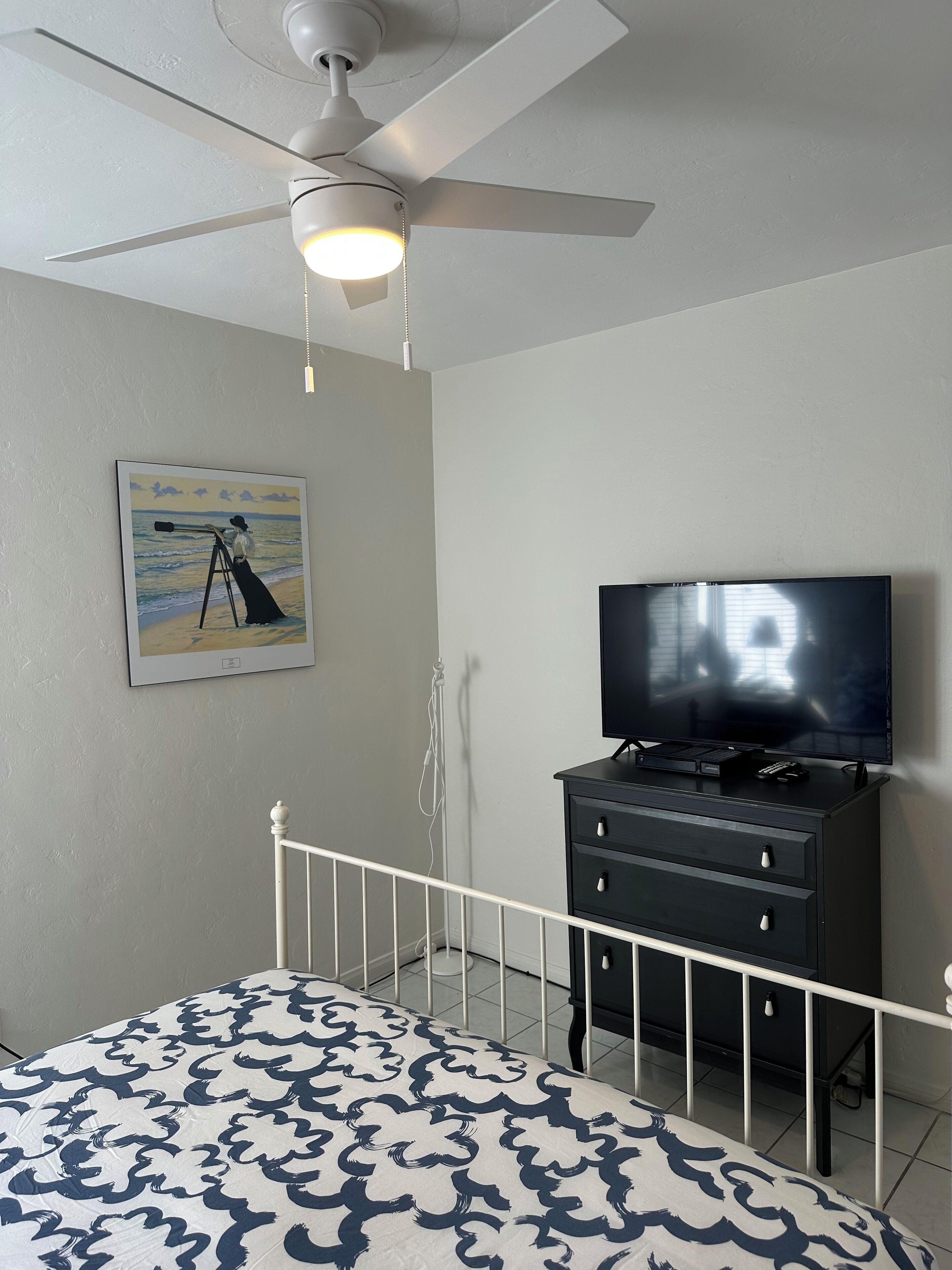 A bedroom with a bed, a ceiling fan, a television on a dresser, and a painting on the wall.