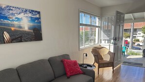 A living room with a gray sofa, a wicker chair, a black rug, and a painting of a beach scene. - Newly refreshed  Beach Apartment..Steps to the sand! (San Diego)