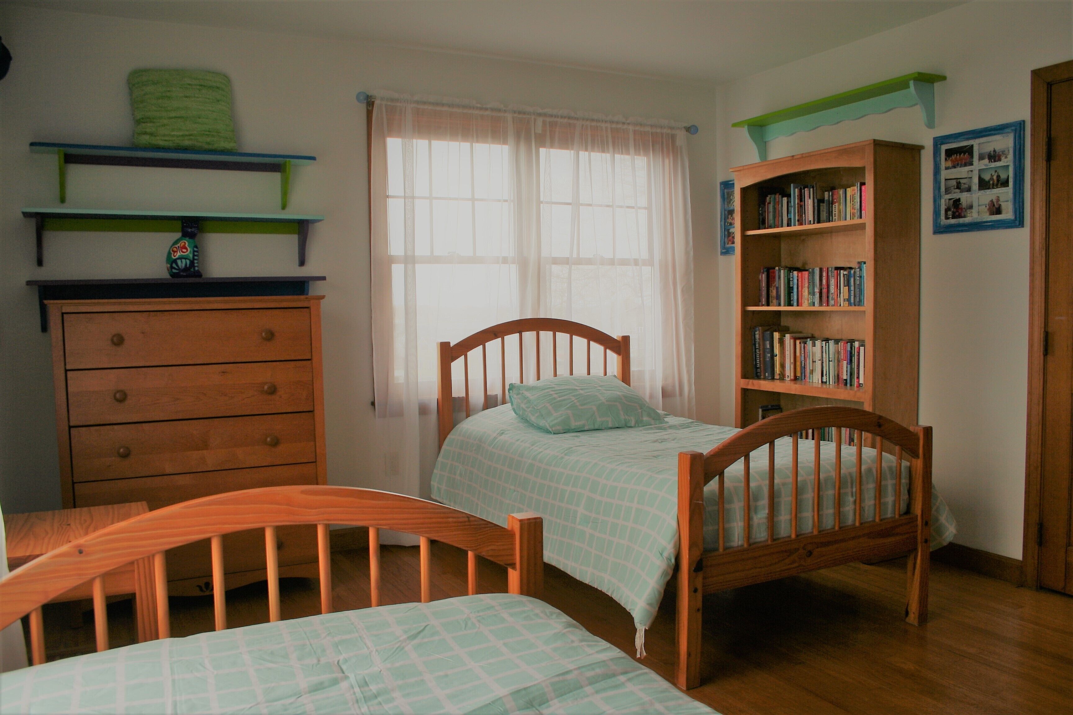 A bedroom with a wooden bed, a wooden dresser, a bookshelf, and a window with sheer curtains.