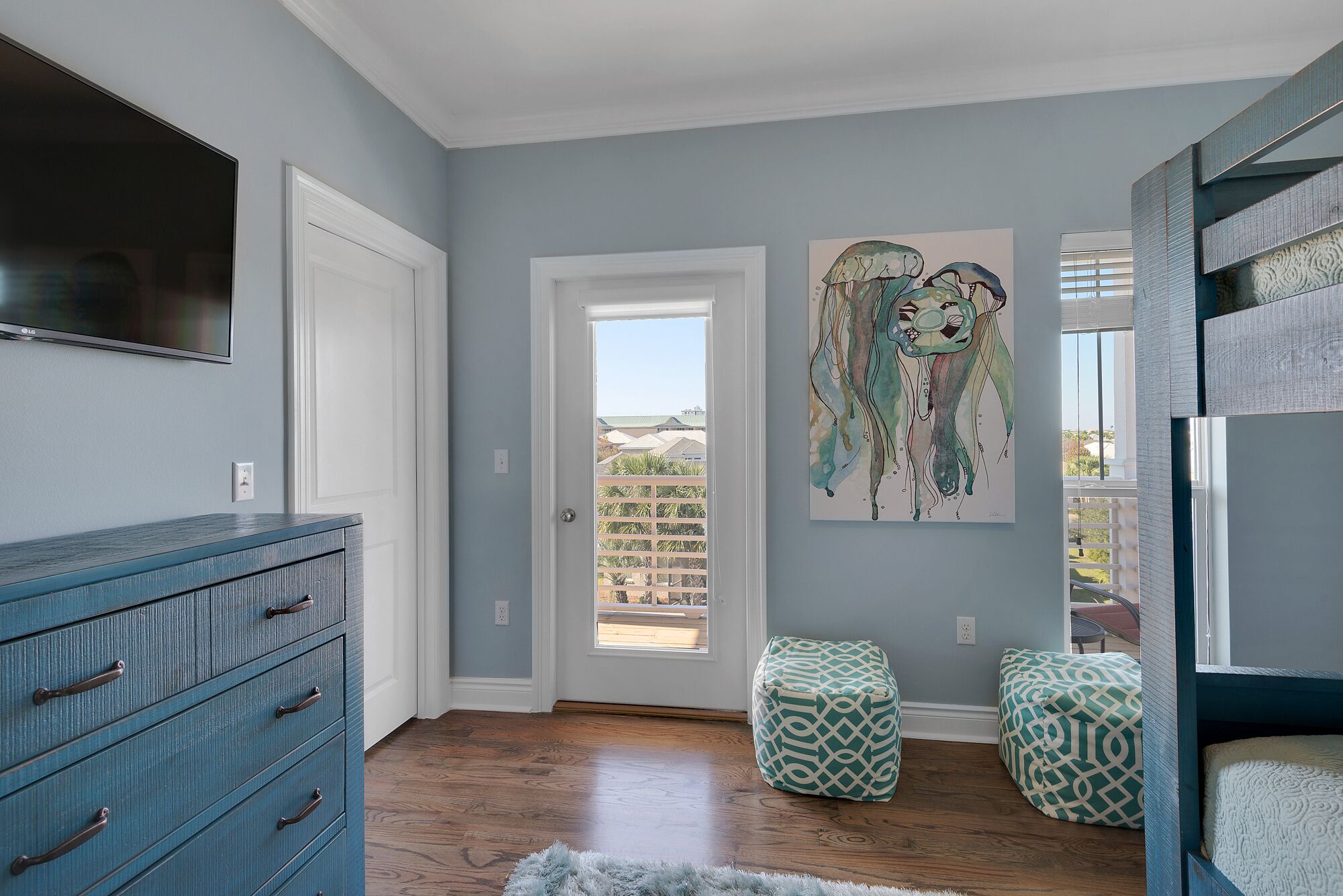 A room with a blue wall, a wooden floor, a blue dresser, two patterned ottomans, a painting, and a door leading outside.