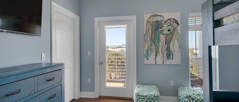 A room with a blue wall, a wooden floor, a blue dresser, two patterned ottomans, a painting, and a door leading outside.