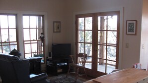 A living room with a TV, a sofa, a chair, and a wooden table. There are large windows and a door leading outside.