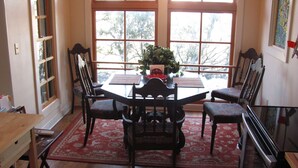 A dining area with a table and chairs, a red patterned rug, and large windows with a view of trees.