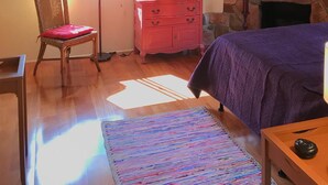 A cozy room with a wooden ceiling, a stone fireplace, a television on a pink dresser, a wicker chair with a red cushion, and a purple bedspread.