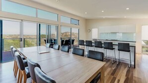 Un comedor amplio con una larga mesa y sillas, una cocina con armarios blancos, y ventanas grandes que ofrecen vistas del paisaje.