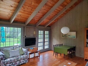 A room with a wooden ceiling, a sofa, a dining table with chairs, a television, and a window with a view of greenery. - Prud'homme Cottage: bright, private architectural cottage on Mitchell Cove (Bernard)