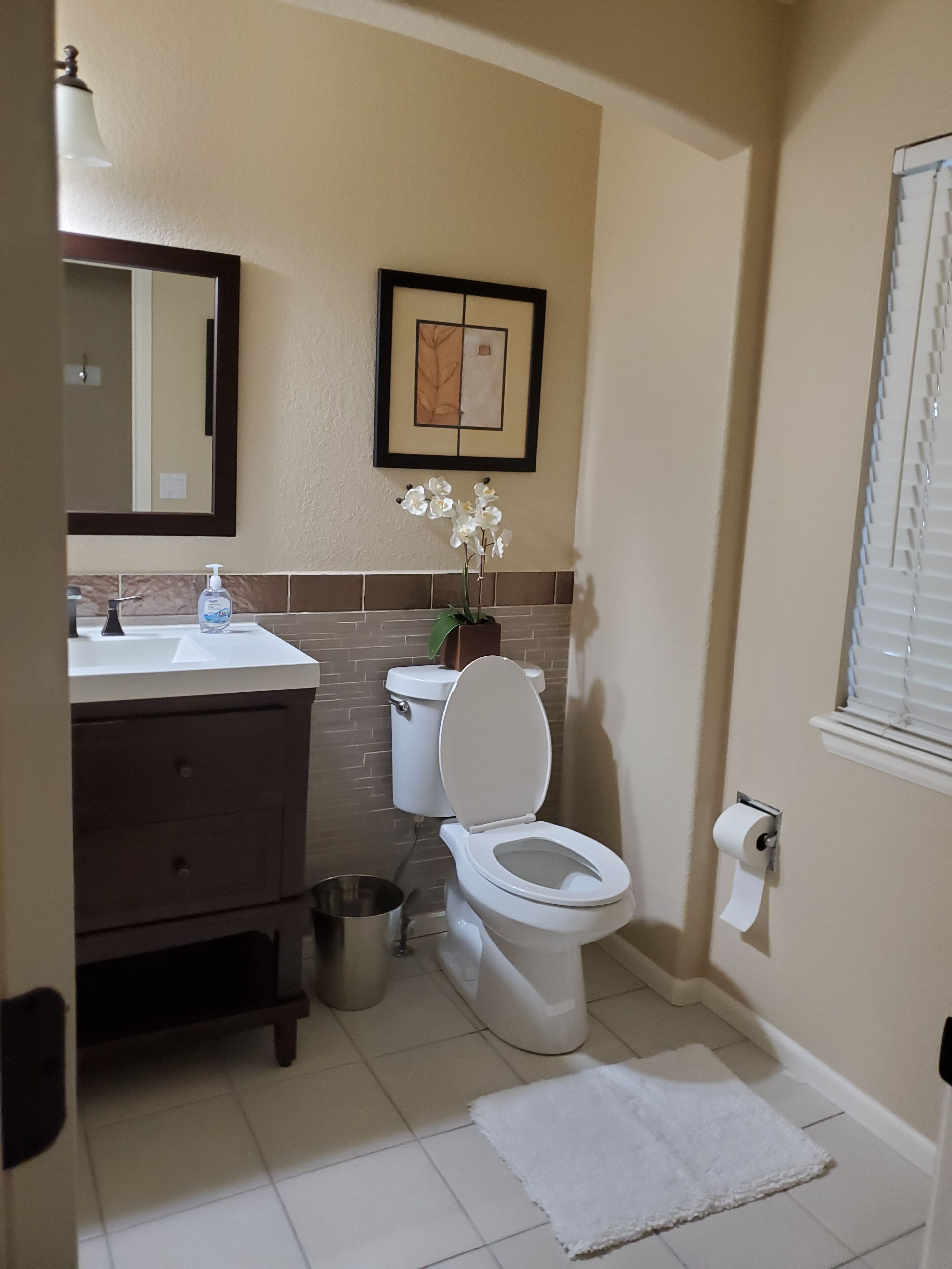 Une salle de bain avec des toilettes, un lavabo et un miroir. Il y a un tableau encadré au mur et une petite poubelle.