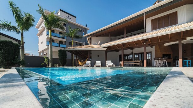 Una piscina con agua azul cristalina, rodeada por un solárium con baldosas. Hay sillones de descanso y una zona de estar cubierta con mesas.