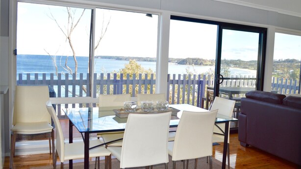 Un espace repas avec une table en verre et des chaises blanches, de grandes fenêtres offrant une vue sur la mer et une terrasse en bois.