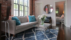 A living room with a sofa, coffee table, and a mirror. There is a brick wall and a window with blinds.