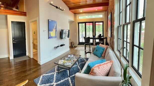 A spacious living area with a wooden ceiling, large windows, a sofa with colorful cushions, and a dining area with a table and chairs.