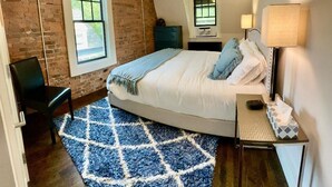 A bedroom with a large bed, a blue and white patterned rug, a brick wall, a window, a desk with a lamp, and a chair.