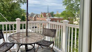A balcony with a round table and two chairs, a red umbrella, and a view of a church steeple in the background.